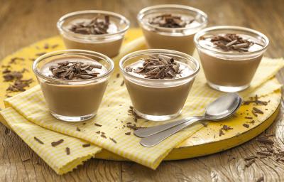 Fotografia em tons de amarelo em uma bancada de madeira, uma tábua amarela redonda com um paninho amarelo xadrez e vários copinhos pequenos de vidro com a mousse de chocolate ao leite. E raspas de chocolate por cima para decorar.