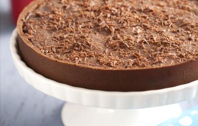 Fotografia em tons de branco e chocolate, ao centro sobre boleira branca uma torta de achocolatado com raspas de chocolate, ao lado guardanapo azul com bolinhas brancas, copo vermelho com talheres e pratos ao fundo, tudo sobre bancada em tons de cinza. Fotografia em tons de branco e chocolate, ao centro sobre boleira branca uma torta de achocolatado com raspas de chocolate, ao lado guardanapo azul com bolinhas brancas, copo vermelho com talheres e pratos ao fundo, tudo sobre bancada em tons de cinza.