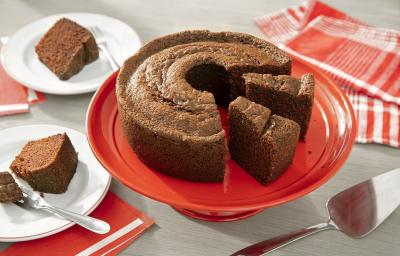 Fotografia em tons de marrom em uma bancada de madeira de cor cinza. Ao centro, uma boleira vermelha contendo o bolo de chocolate. Ao fundo, há um pano vermelho listrado e ao lado, há 2 pires com 2 fatias de bolo. Fotografia em tons de marrom em uma bancada de madeira de cor cinza. Ao centro, uma boleira vermelha contendo o bolo de chocolate. Ao fundo, há um pano vermelho listrado e ao lado, há 2 pires com 2 fatias de bolo.