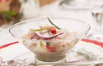 Fotografia em tons de branco e vermelho de uma mesa branca com um prato vermelho, sobre ele um prato branco florido, uma colher prata, e uma taça com pedaços de peixe, cebola roxa, pimenta e cebolinha. Ao fundo um arranjo de flores. Fotografia em tons de branco e vermelho de uma mesa branca com um prato vermelho, sobre ele um prato branco florido, uma colher prata, e uma taça com pedaços de peixe, cebola roxa, pimenta e cebolinha. Ao fundo um arranjo de flores.