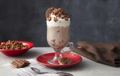 Foto de uma mesa cinza. Ao centro há um prato vermelho com uma caneca alta com a bebida de Chocolate Quente, decorada com Chantilly e o crumble. Na base há um biscoito Calipso e mais dois na mesa. Ao fundo há um prato com mais crumble. Foto de uma mesa cinza. Ao centro há um prato vermelho com uma caneca alta com a bebida de Chocolate Quente, decorada com Chantilly e o crumble. Na base há um biscoito Calipso e mais dois na mesa. Ao fundo há um prato com mais crumble.