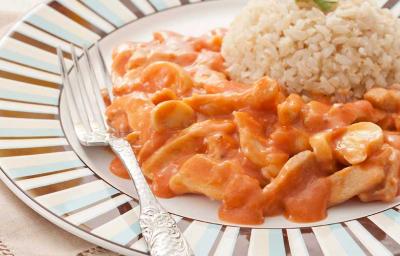 Fotografia em tons de marrom em uma bancada com uma toalha bege, um prato branco raso grande com detalhes listrados em azul e marrom, com o strogonoff de frango com molho e arroz integral.