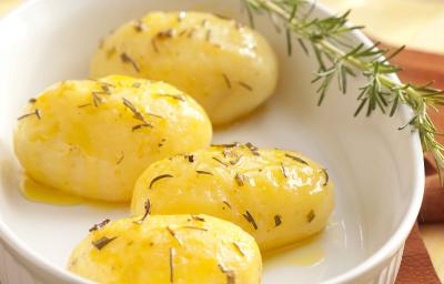 Fotografia em tons de branco e amarelo de uma bancada de madeira clara, com um pano marrom, um refratário branco oval com 4 batatas dentro, com azeite e alecrim e um ramo de alecrim. Fotografia em tons de branco e amarelo de uma bancada de madeira clara, com um pano marrom, um refratário branco oval com 4 batatas dentro, com azeite e alecrim e um ramo de alecrim.