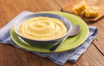Fotografia em tons de verde e azul em uma bancada de madeira clara, com um paninho azul, um prato verde e outro azul fundo em cima com o creme de milho. Ao fundo, uma tábua de madeira com milhos cortados. Fotografia em tons de verde e azul em uma bancada de madeira clara, com um paninho azul, um prato verde e outro azul fundo em cima com o creme de milho. Ao fundo, uma tábua de madeira com milhos cortados.