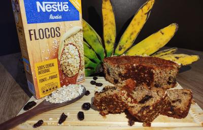 Foto aproximada de um bolo em tom marrom, cortado em pedaços, ao lado esquerdo há uma caixa de aveia em flocos nestlé, uma colher com um pouco de aveia e ao fundo há um cacho de bananas