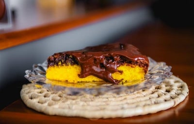 Fotografia de uma fatia de bolo de cenoura com brownie por cima e uma calda de chocolate brilhante para finalizar. A fatia está em um prato de vidro pequeno, apoiado em um apoio raso de bambu de tom claro. Fotografia de uma fatia de bolo de cenoura com brownie por cima e uma calda de chocolate brilhante para finalizar. A fatia está em um prato de vidro pequeno, apoiado em um apoio raso de bambu de tom claro.