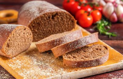 Fotografia de uma tábua média de madeira sobre uma mesa de madeira escura. Sobre a tábua tem uma receita de pão australiano com três fatias cortadas e sobrepostas ao lado de dois pedaços maiores do pão.