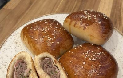 Fotografia de três hambúrgueres de forno recheados com presunto e queijo sobre um prato branco apoiado em uma tábua de madeira. Ao lado dos hambúrgueres tem mais um, mas que está aberto ao meio. Fotografia de três hambúrgueres de forno recheados com presunto e queijo sobre um prato branco apoiado em uma tábua de madeira. Ao lado dos hambúrgueres tem mais um, mas que está aberto ao meio.