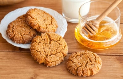 Em cima de uma mesa, prato branco com biscoitos crocantes, decorados com granulado de açúcar cristal. Ao lado do prato, pote com mel.
