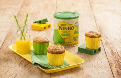 Fotografia em tons de amarelo e verde em uma bancada de madeira clara, um recipiente amarelo com dois cupcakes de maçã e aveia em forminhas amarela e verde. Ao lado, brinquedinhos infantis, uma lata de Ninho Fases e mais um cupacke. Fotografia em tons de amarelo e verde em uma bancada de madeira clara, um recipiente amarelo com dois cupcakes de maçã e aveia em forminhas amarela e verde. Ao lado, brinquedinhos infantis, uma lata de Ninho Fases e mais um cupacke.