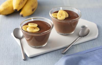 Fotografia em tons de azul e marrom de uma bancada com um paninho azul, duas tacinhas transparentes com o creme de chocolate e rodelas de banana decorando, ao lado duas colheres pratas e ao fundo 3 bananas. Fotografia em tons de azul e marrom de uma bancada com um paninho azul, duas tacinhas transparentes com o creme de chocolate e rodelas de banana decorando, ao lado duas colheres pratas e ao fundo 3 bananas.