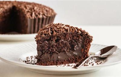 Fotografia em tons claros de uma fatia grande de bolo de chocolate molhadinho com brigadeiro e granulado por cima ao lado de uma colher, sobre um prato raso branco. Fotografia em tons claros de uma fatia grande de bolo de chocolate molhadinho com brigadeiro e granulado por cima ao lado de uma colher, sobre um prato raso branco.