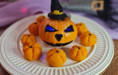 Fotografia em tons de laranja, em uma bancada de madeira, um prato branco ao centro com os beijinhos em formato de abóbora. No centro, uma abóbora pequena com desenho de Halloween e um chapeuzinho de bruxa ao fundo da foto. Fotografia em tons de laranja, em uma bancada de madeira, um prato branco ao centro com os beijinhos em formato de abóbora. No centro, uma abóbora pequena com desenho de Halloween e um chapeuzinho de bruxa ao fundo da foto.
