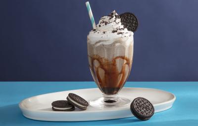 Foto da receita de Milkshake de negresco. Observa-se um copo de milkshake de vidro transparente decorado com calda e com a bebida dentro e topping de chantilly e farofa de biscoito, com um canudo azul e branco espetado