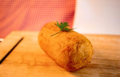 Fotografia de um croquete frito de carne moída sobre uma tábua de madeira. Tem um pequeno ramo de salsinha decorando o croquete. Fotografia de um croquete frito de carne moída sobre uma tábua de madeira. Tem um pequeno ramo de salsinha decorando o croquete.