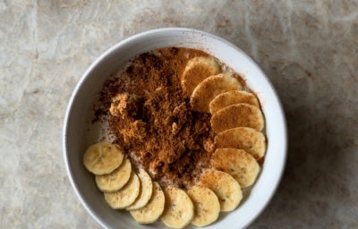 Fotografia de um pote branco com mingua de aveia dentro e rodelas de banana com canela decorando