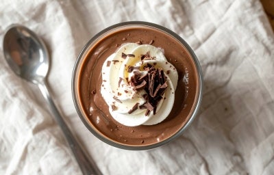 Fotografia vista de ciam de um recipiente de vidro com a sobremesa, finalziada com ganache de chocolate, chantilly e raspas de chocolate. Ao lado tem uma colher de sobremesa, e o recipiente está sobre um pano de tom claro com texturas.
