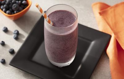 Fotografia em tons de preto, laranja e branco de uma bancada de madeira, sobre ela um prato quadrado preto com um copo de smoothie. Ao lado um paninho laranja e um recipiente laranja com mirtilo. Fotografia em tons de preto, laranja e branco de uma bancada de madeira, sobre ela um prato quadrado preto com um copo de smoothie. Ao lado um paninho laranja e um recipiente laranja com mirtilo.