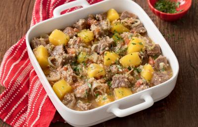 Fotografia em tons de marrom e branco uma bancada vista de cima, ao centro um recipiente quadrado com alças cor branco, dentro dele cubos de carne e pedaços de Mandioquinha e Quinoa. Ao lado um pano listrado na cor vermelho e branco. Fotografia em tons de marrom e branco uma bancada vista de cima, ao centro um recipiente quadrado com alças cor branco, dentro dele cubos de carne e pedaços de Mandioquinha e Quinoa. Ao lado um pano listrado na cor vermelho e branco.