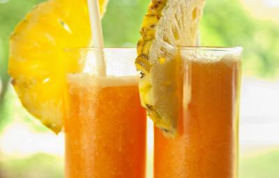 Fotografia de dois copos de vidro grandes com suco de laranja, abacaxi e mel. Em cada copo, tem metade de uma fatia de abacaxi e tem também um canudo. Ao fundo tem uma paisagem com de natureza com árvores. Fotografia de dois copos de vidro grandes com suco de laranja, abacaxi e mel. Em cada copo, tem metade de uma fatia de abacaxi e tem também um canudo. Ao fundo tem uma paisagem com de natureza com árvores.