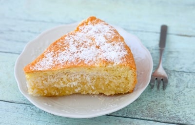 Fotografia de uma fatia de bolo de coco em um prato decorativo, com cobertura de açúcar de confeiteiro e uma crosta dourada, sobre uma superfície de madeira azul clara, acompanhada de um garfo metálico.