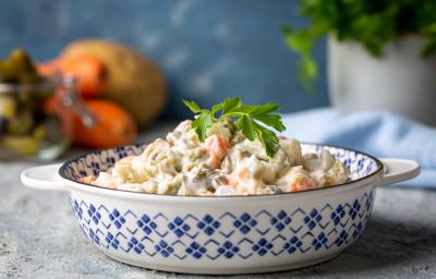 Fotografia de uma salada de maionese que está em um recipiente fundo com desenhos azuis. Tem um ramo de salsinha apoiado na salada. O recipiente está apoiado em uma bancada de textura parecida com gesso, ao fundo tem frutas. Fotografia de uma salada de maionese que está em um recipiente fundo com desenhos azuis. Tem um ramo de salsinha apoiado na salada. O recipiente está apoiado em uma bancada de textura parecida com gesso, ao fundo tem frutas.