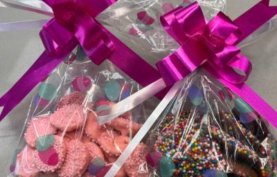 Imagem de dois saquinhos plásticos com laço grande cor de rosa sobre cada um deles, apoiados em um fundo claro. Dentro dos saquinhos, pequenas porções de chocolate cor de rosa e marrom com confeitos coloridos.