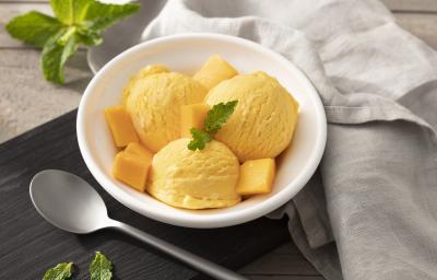 Fotografia em tons de branco, preto e cinza de uma bancada cinza com um paninho preto, sobre ele um recipiente branco redondo com sorvete de manga. Ao lado um paninho cinza e uma colher. Fotografia em tons de branco, preto e cinza de uma bancada cinza com um paninho preto, sobre ele um recipiente branco redondo com sorvete de manga. Ao lado um paninho cinza e uma colher.