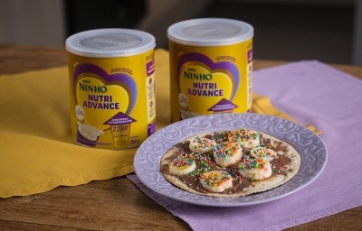 Fotografia de dois pano, roxo e amarelo, em uma mesa de madeira. Sobre esses panos tem duas latas de NINHO Nutri Advance ao lado de um prato roxo claro com uma mini pizza com chocolate e banana.