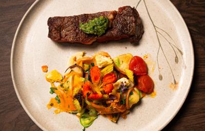 Fotografia vista de cima de um bife ancho com pesto de cerefólio e salada de cogumelos ao lado. A receita está em um prato grande, branco e raso, o qual está sobre uma mesa de madeira.