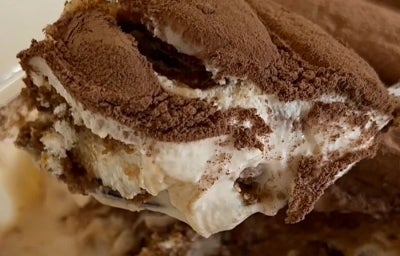 Fotografia em tons claros de um pedaço de tiramisù carregado por uma colher. No fundo tem o restante da receita em um recipiente refratário de vidro retangular.