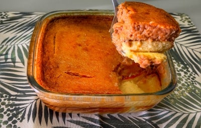 Fotografia de uma travessa quadrada de vidro sobre um pano de mesa brnaco com desenhos pretos. Dentro da travessa tem uma sobremesa de Goiabada Nordestina Cremosa com Queijo Coalho em camadas.