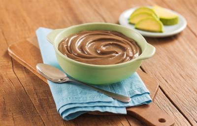 Fotografia em tons de marrom, em uma bancada de madeira de cor marrom. No centro, há uma tábua de madeira com um pano azul claro. Em cima, há um recipiente contendo o mousse com uma colher ao lado. Ao fundo, um pires contendo algumas fatias de abacate.
