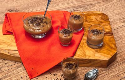 Fotografia em tons de laranja, em uma bancada de madeira, uma tábua de madeira, um pano laranja e 5 recipientes de vidro em cima dele, com os brigadeiros de colher com chocolate granulado em cima do pano e tábua.