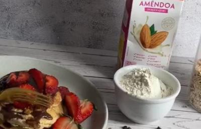 Foto em tons de cinza da receita de panqueca plant-based servida com calda e morangos sobre um prato de porcelana branco. Ao lado, há uma xícara com farinha de trigo e um bowl branco com morangos, além de uma caixa de bebida vegetal nature's heart