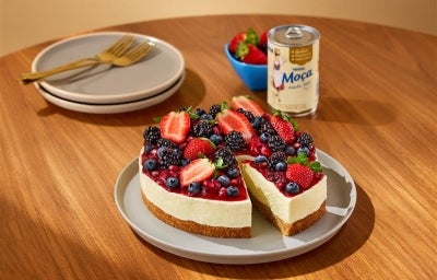 Fotografia mostra uma fatia de cheesecake de frutas vermelha sendo retirada e a lata de leite moça.