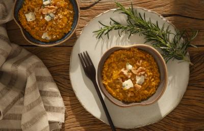 Foto em tons de laranja da receita de risoto de abóbora com gorgonzola servida em um bowl de cerâmica cinza sobre uma base de porcelana branca com um garfo ao lado além de um ramo de alecrim