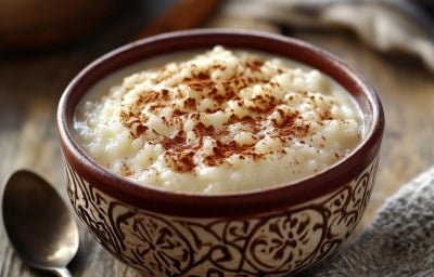 Em cima de uma mesa, tigela marrom com arroz doce cremoso, com uma textura suave e aveludada, decorado com canela em pó.