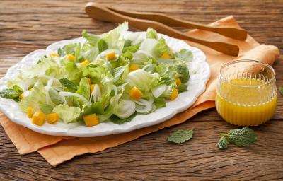 Fotografia em tons de marrom em uma bancada de madeira com um pano laranja e um prato branco oval com as hortaliças. Ao lado, um pegador de salada de madeira e um copo de vidro com suco de laranja.