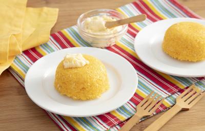 Foto em tons de amarelo da receita de cuscuz de micro-ondas com queijo servida em duas porções sobre pratos brancos de porcelana em cima de uma toalhinha colorida com garfos de madeira e um vidro com queijo ao lado