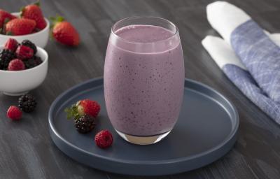 Foto em tons de roxo da receita de shake de frutas vermelhas com chia servida em um corpo largo sobre uma base de porcelana azul escura circular com frutas vermelhas ao lado. Ao fundo, há dois recipientes com frutas vermelhas e um pano azul.