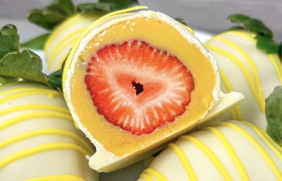 Fotografia de um morango envolto em um brigadeiro de maracujá, coberto com uma casquinha de chocolate branco