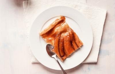 Fotografia vista de cima de uma torta de banana com bananas caramelizadas por cima em um prato de sobremesa branco, e uma colher ao lado. A sobremesa está em cima de um pano branco, que está sobre uma mesa branca.