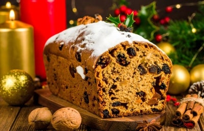 Fotografia de um bolo de frutas festivo, exibido sobre uma superfície de madeira, com textura densa e decorado com açúcar em pó. Ao redor, velas acesas, nozes e canela.