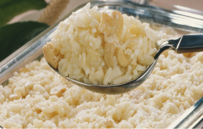 Fotografia em tons de branco e bege, ao centro colher de servir com arroz e cogumelos sobre refratário com o mesmo arroz, ao fundo folhas verdes como decoração. Fotografia em tons de branco e bege, ao centro colher de servir com arroz e cogumelos sobre refratário com o mesmo arroz, ao fundo folhas verdes como decoração.