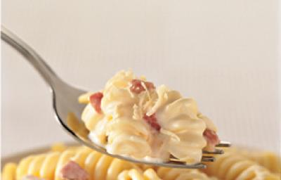 Fotografia em tons de amarelo de um garfo prateado com uma porção de macarrão parafuso cremoso com bacon sobre um pote com macarrão.