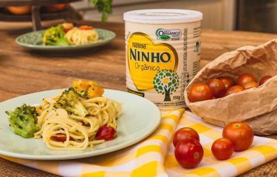 Fotografia em tons de amarelo de uma bancada de madeira com um pano xadrez amarelo e branco de um prato verde claro com o macarrão, brócolis, abóbora e tomate cereja. Ao fundo uma lata de NINHO. Fotografia em tons de amarelo de uma bancada de madeira com um pano xadrez amarelo e branco de um prato verde claro com o macarrão, brócolis, abóbora e tomate cereja. Ao fundo uma lata de NINHO.