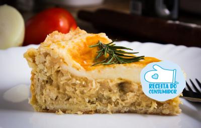 Fotografia em tons de amarelo, em uma bancada de madeira de cor marrom. Ao centro, um prato branco contendo uma fatia da torta de frango. Ao fundo, há um tomate e um pedaço de cebola. Fotografia em tons de amarelo, em uma bancada de madeira de cor marrom. Ao centro, um prato branco contendo uma fatia da torta de frango. Ao fundo, há um tomate e um pedaço de cebola.