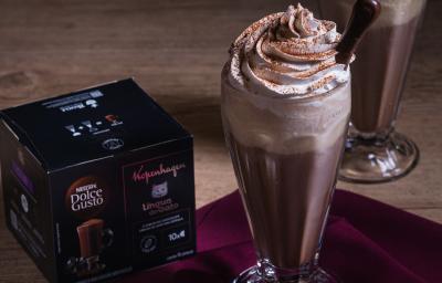 Fotografia em tons escuros de um copo de vidro de milkshake com uma bebida gelada de kopenhagen dole gusto com chantilly e uma língua de gato apoiado. O copo está ao lado da caixa dolce gusto. Fotografia em tons escuros de um copo de vidro de milkshake com uma bebida gelada de kopenhagen dole gusto com chantilly e uma língua de gato apoiado. O copo está ao lado da caixa dolce gusto.