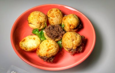 Fotografia de um prato vermelho sobre uma mesa branca. Dentro do prato tem seis pães de queijo recheados com carne de panela.